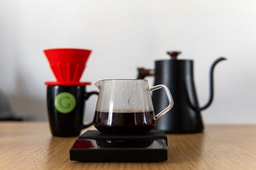 A minimalist pour-over coffee setup featuring a glass carafe on a scale, with a red coffee filter and black kettle.