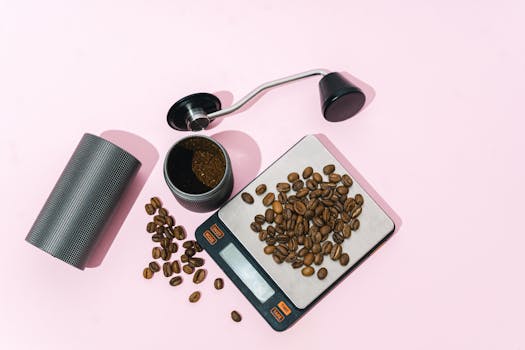 Top view of coffee beans on a digital scale with a grinder on a pink background.